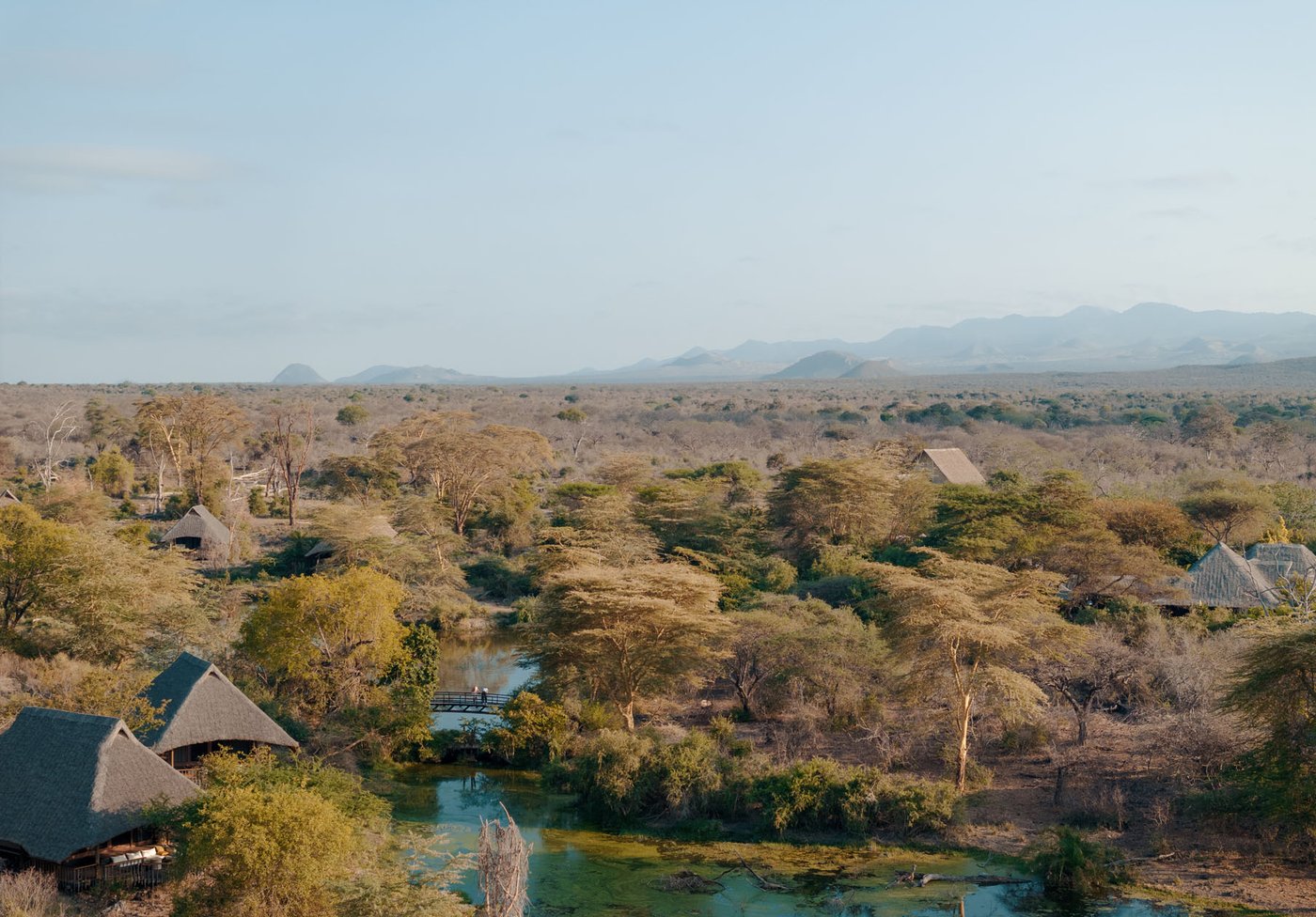 Thatched suites among acacia above the spring-fed pools at Finch Hattons, with the hills of Tsavo West behind.