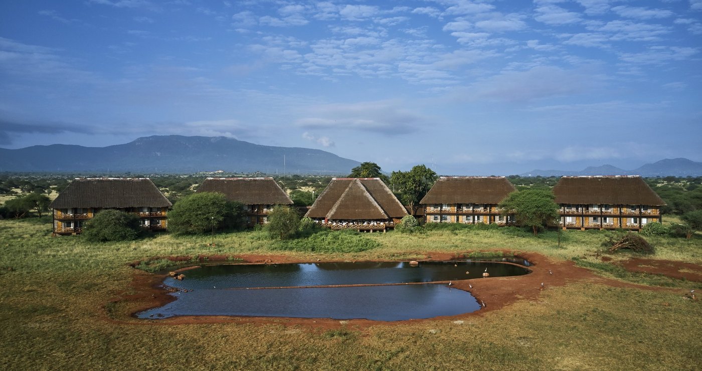 Thatched lodge buildings beside a round waterhole with hills behind at Ngutuni Safari Lodge in Tsavo