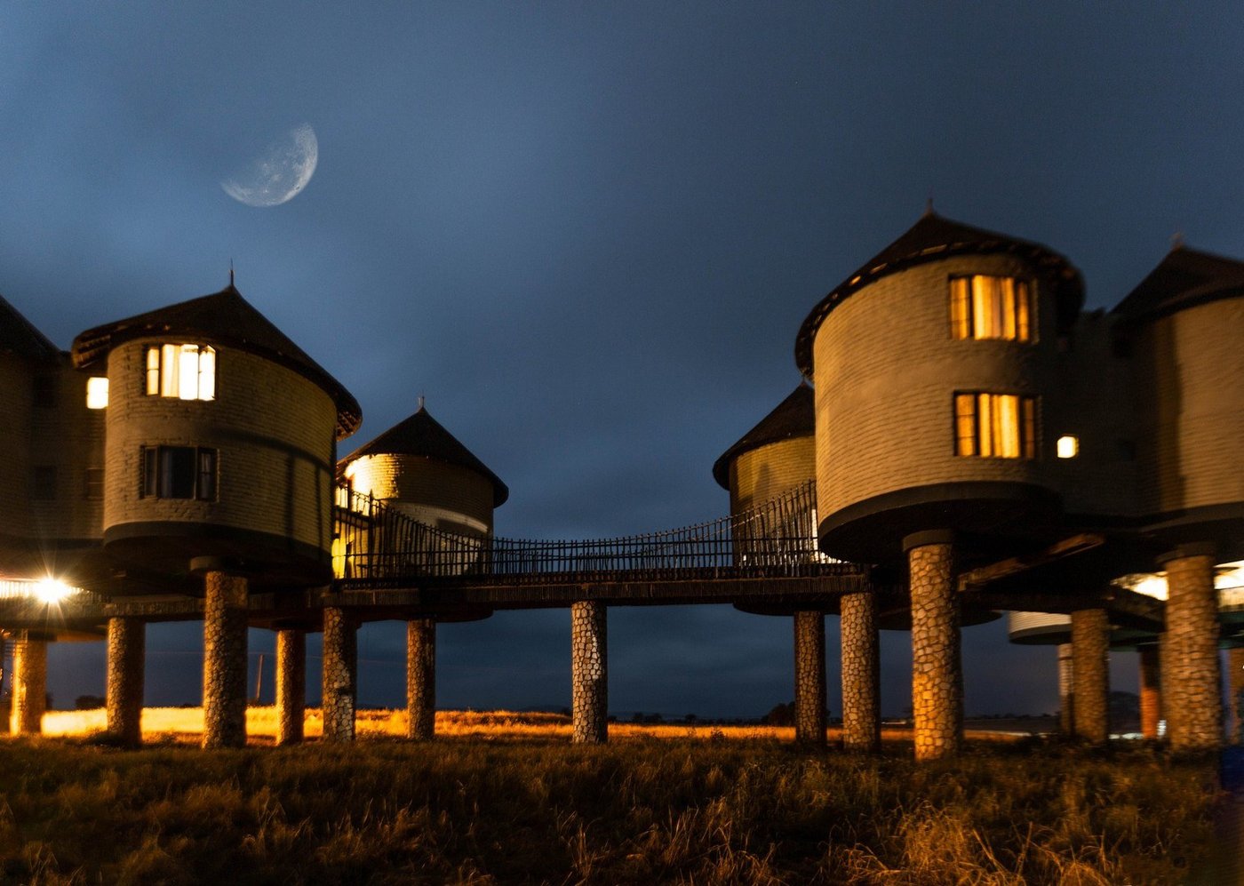 Stilted rondavels lit at dusk under a crescent moon at Salt Lick Safari Lodge, Tsavo